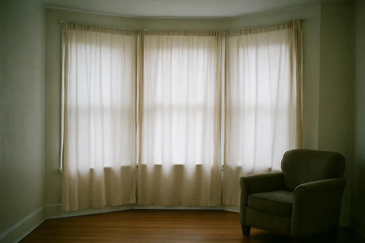 A three-sided bay window in a bright living room with floor-length neutral curtains framing each angled section, warm daylight coming through.